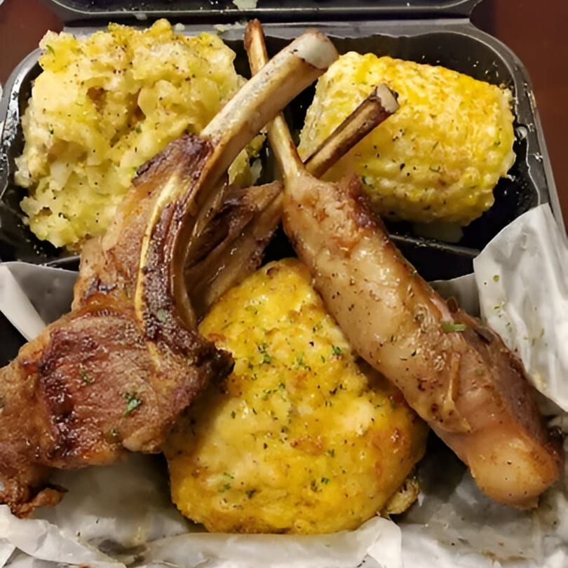Grilled lamb chops, macaroni and cheese, and corn on the cob in a takeout container.