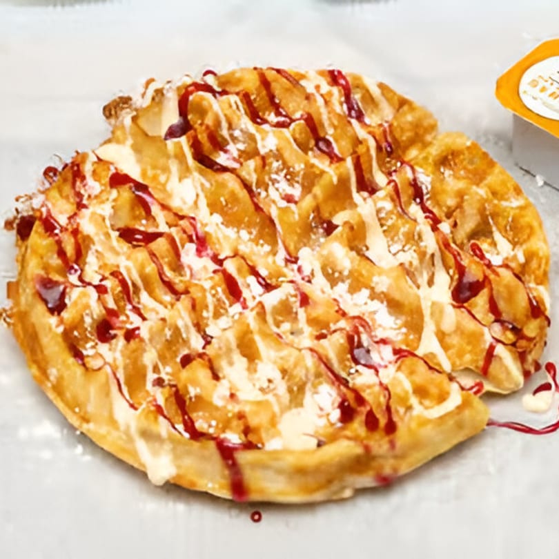 A golden waffle drizzled with white icing and red berry sauce, dusted with powdered sugar.